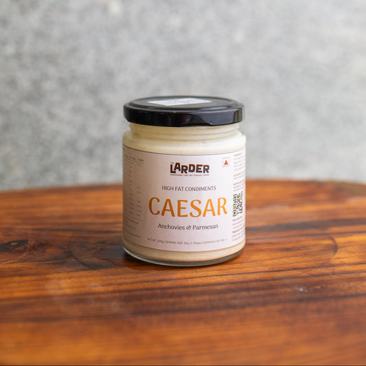 A glass jar of Lärder Caesar high fat condiments, featuring a cream-colored label with orange text and a black lid, sitting on a rustic wooden surface against a textured gray background.