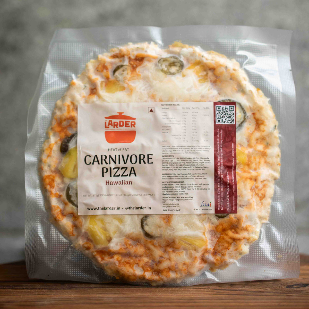 A vacuum-sealed package of The Larder Hawaiian Carnivore Pizza, showing the round pizza with melted cheese, pineapple, and olives inside a clear plastic bag on a wooden surface.