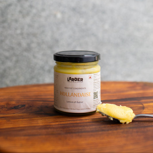 A jar of Larder Hollandaise lemon butter spread sits on a wooden board with a spoonful of the yellow spread in front, set against a textured gray background.