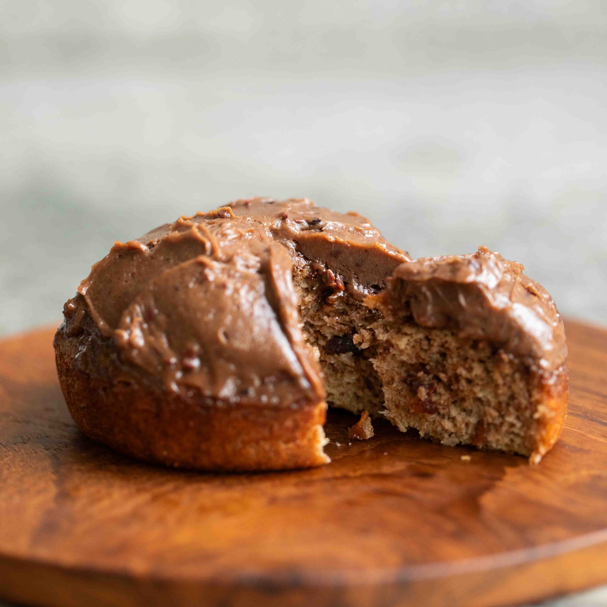 A close-up of The Larder Keto chocolate cake with thick chocolate frosting, with a slice cut out to show the moist interior. The cake sits on a round wooden serving board against a softly blurred light gray background.
