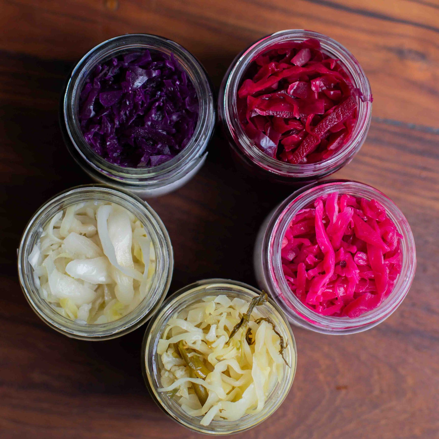 Five jars of different types of The Larder's variety of sauerkraut on a wooden surface